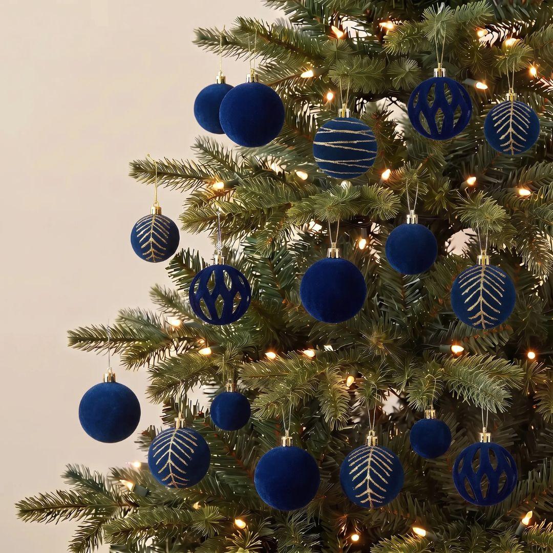 A set of deep blue velvet Christmas tree ornaments with gold metallic patterns, hanging on a green Christmas tree with warm white lights against a neutral beige background.