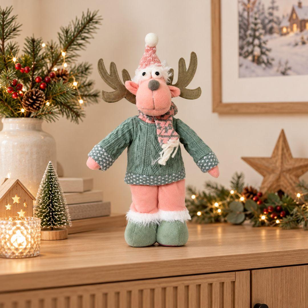 Noel Christmas Plush Design B, 43cm tall pink reindeer decoration featuring brown antlers, sage green knitted sweater, patterned scarf, pink Santa hat and green plush boots, displayed on a wooden sideboard.