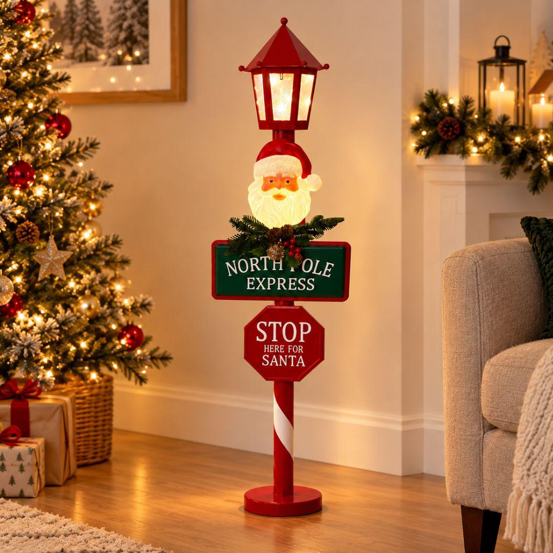 North Pole Express LED Christmas Lamp Post 90cm with illuminated Santa Claus face, glowing red lantern and festive signposts on beige background