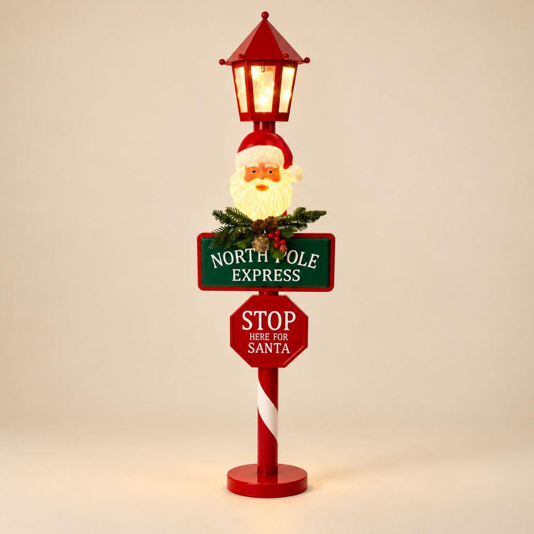 North Pole Express LED Christmas Lamp Post 90cm with illuminated Santa Claus face, glowing red lantern and festive signposts on beige background
