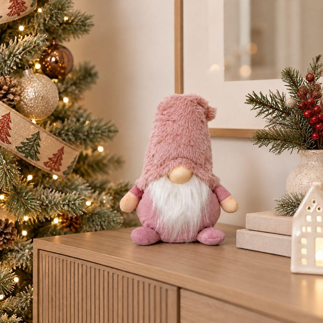 Quillby Gnome Design V, 22cm tall blush pink plush Christmas gnome featuring an oversized fluffy pink hat, long white beard and soft pink body, displayed on a wooden sideboard beside a decorated Christmas tree.