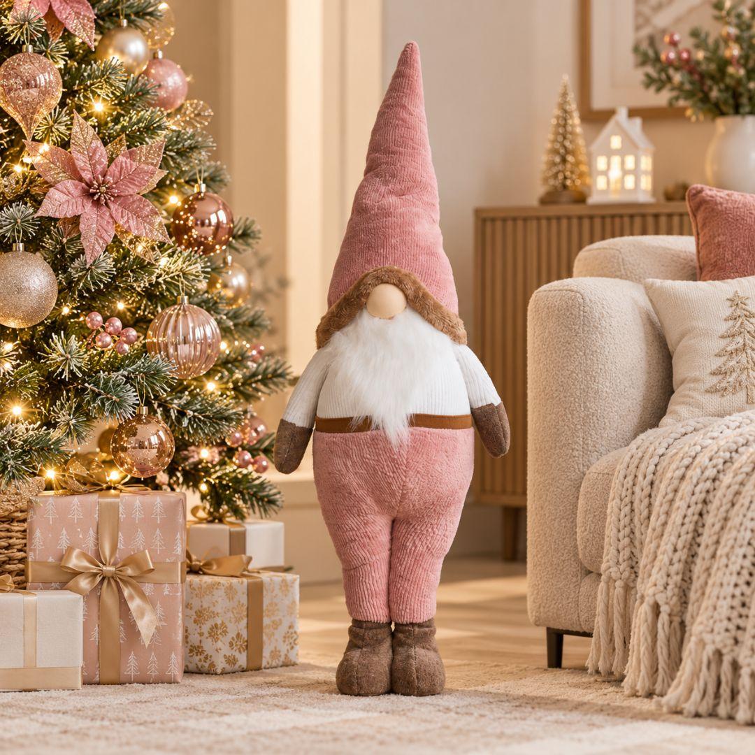 Quillby Gnome Design W, 98cm tall blush pink Christmas gnome featuring an oversized pointed hat, fluffy white beard, pink trousers and brown plush boots, displayed beside a decorated Christmas tree in a cosy living room.