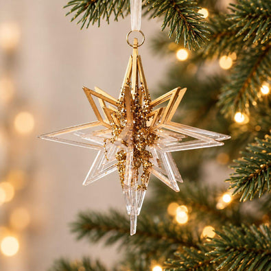 Gold and clear acrylic 3D star Christmas ornament with sparkling gold details hanging on an artificial Christmas tree with warm fairy lights.