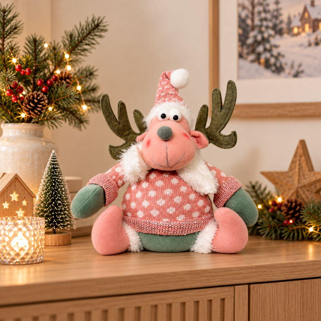 Noel Christmas Plush Design C, 28cm pink sitting reindeer plush decoration featuring brown antlers, pink knitted sweater, festive hat, fluffy white scarf and oversized plush feet, displayed on a wooden sideboard beside Christmas decorations.