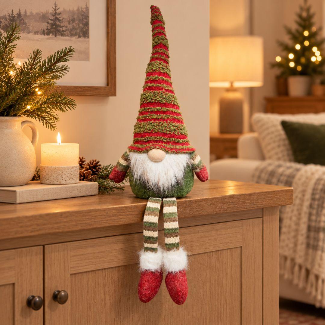 Noel sitting Christmas gnome with tall red and green knitted hat and dangling striped legs displayed on a wooden console in a cosy living room