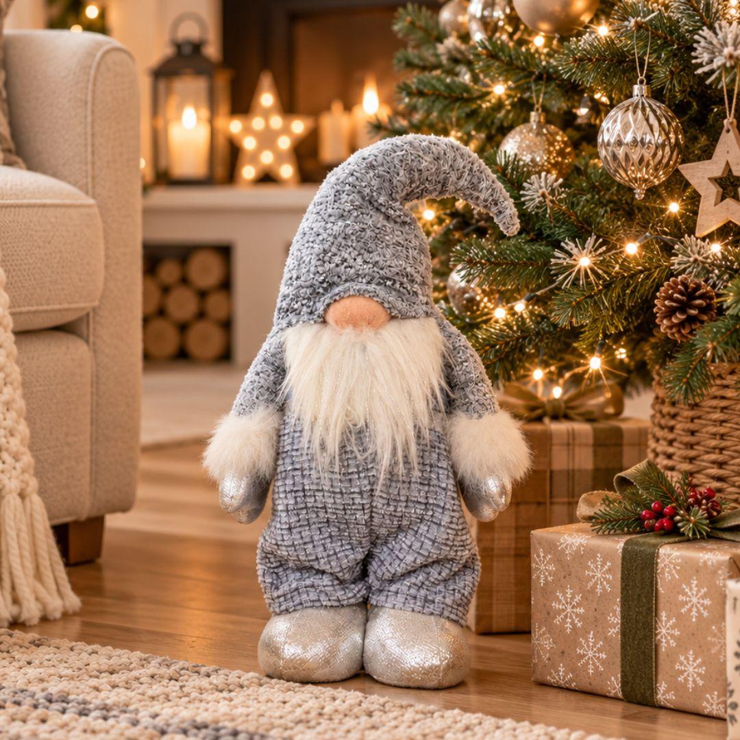 Quillby Gnome Design R, 55cm tall blue plush Christmas gnome with textured floppy hat, fluffy white beard and shimmering silver boots, displayed beside a Christmas tree.
