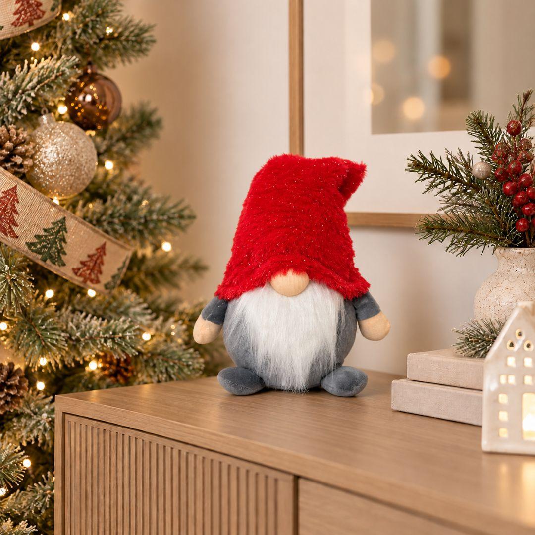 Quillby Gnome Design T, 22cm tall red and grey plush Christmas gnome featuring an oversized fluffy red hat, long white beard and soft grey body, displayed on a wooden sideboard beside a Christmas tree.