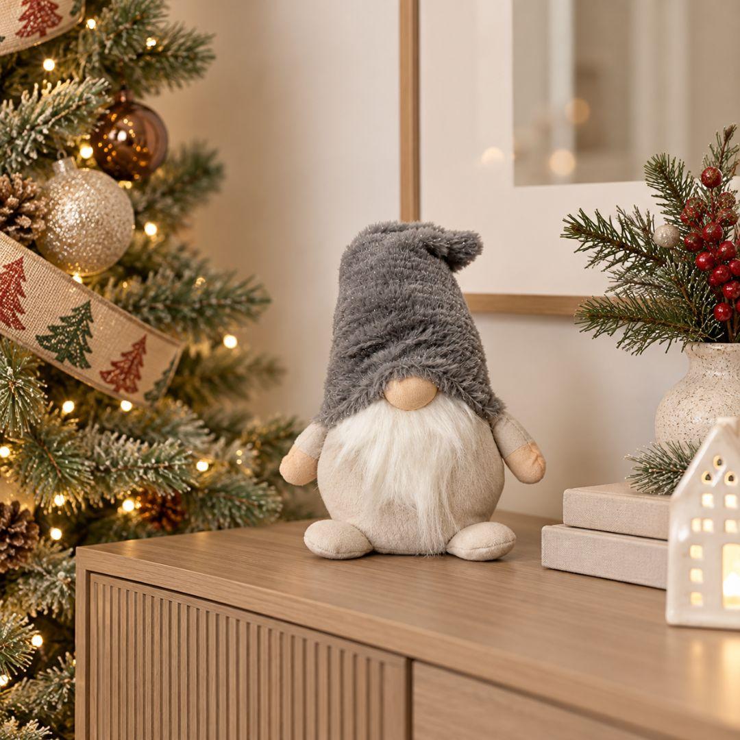 Quillby Gnome Design U, 22cm tall grey and beige plush Christmas gnome featuring an oversized fluffy grey hat, long white beard and soft beige body, displayed on a wooden sideboard beside a decorated Christmas tree.