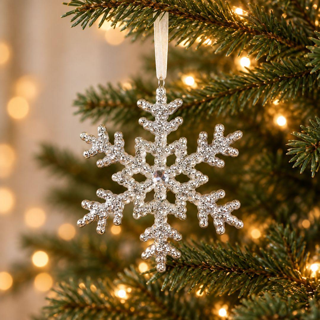 Silver acrylic snowflake Christmas ornament with intricate sparkling details hanging from an artificial Christmas tree decorated with warm LED lights.
