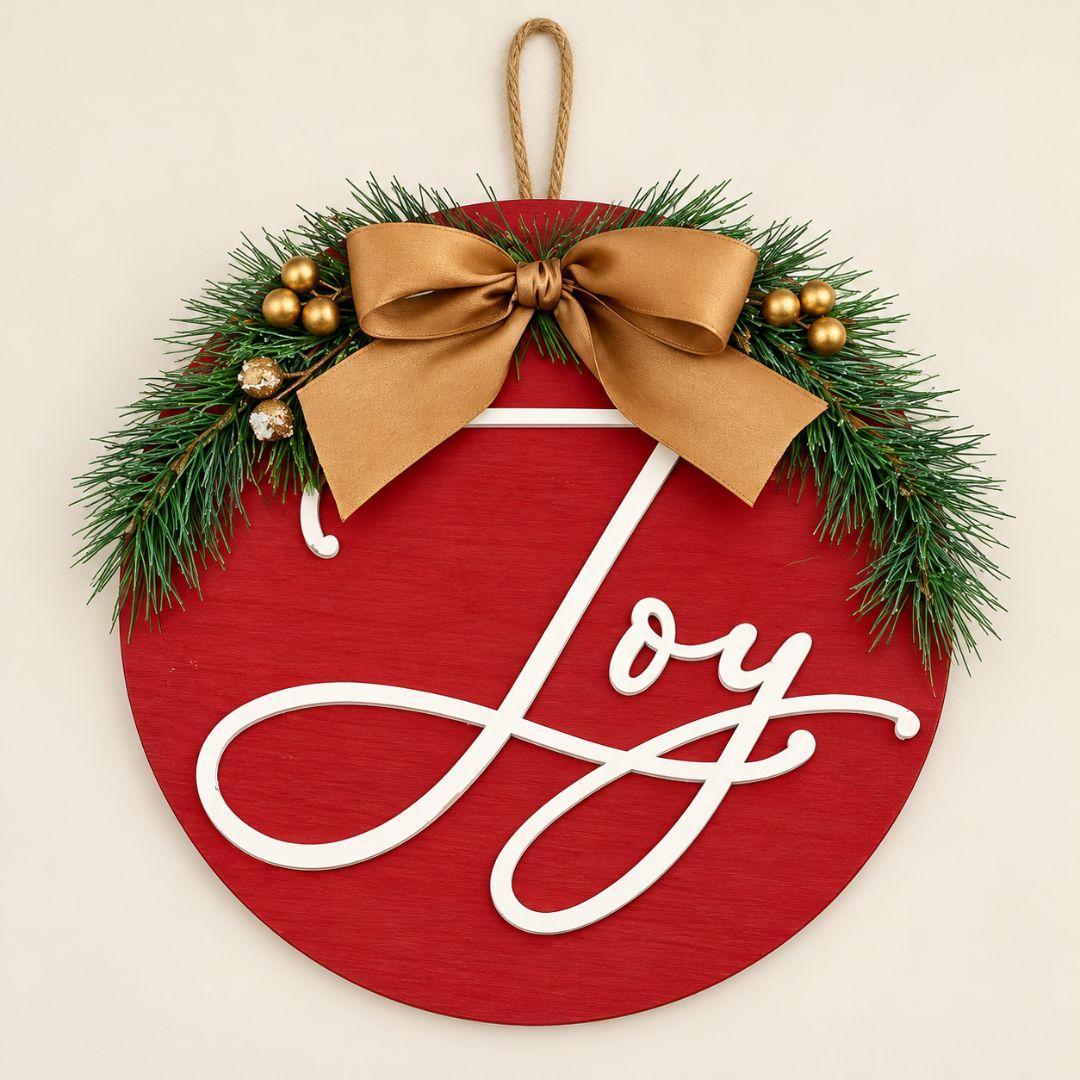 24cm Woodland Joy wooden Christmas wreath featuring white Joy lettering on red wood, pine greenery, gold berries and champagne bow on beige background